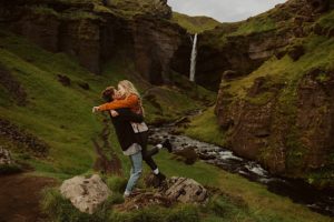 woman with orange jacket hugs fiance before iceland waterfall