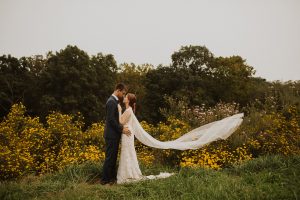 Bride and groom stand in wildflowers with veil flowing in the wind