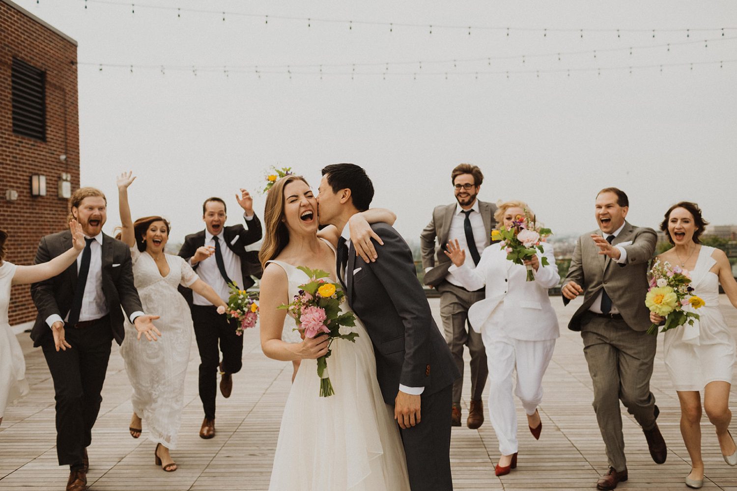Groom kisses bride on cheek with wedding party chasing them
