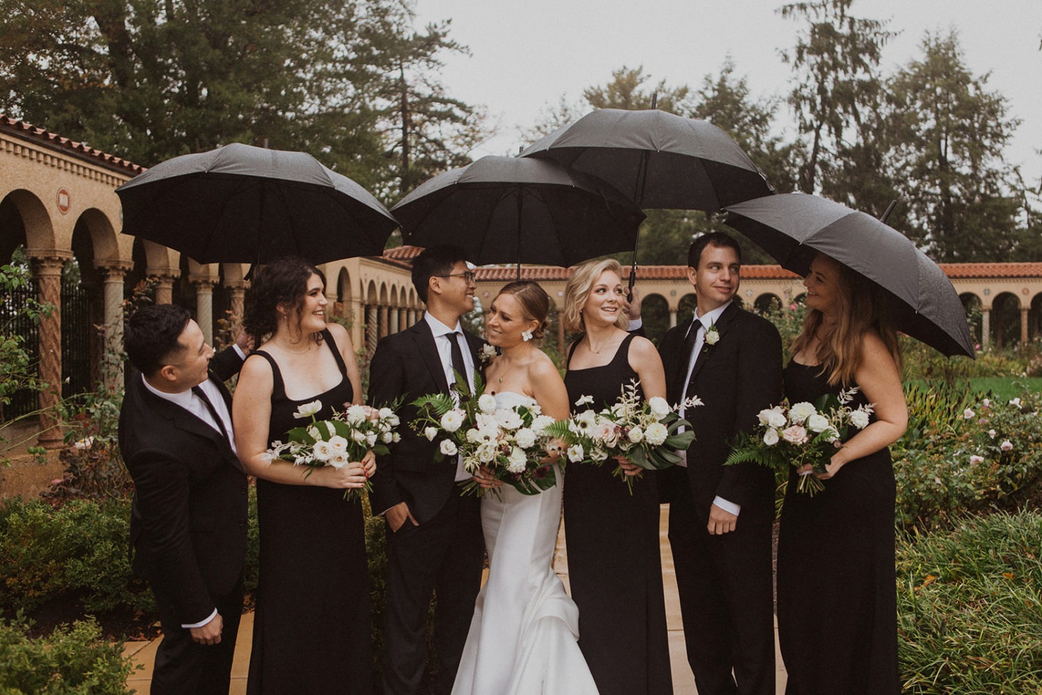 Couple stands under umbrellas with wedding party at intimate wedding