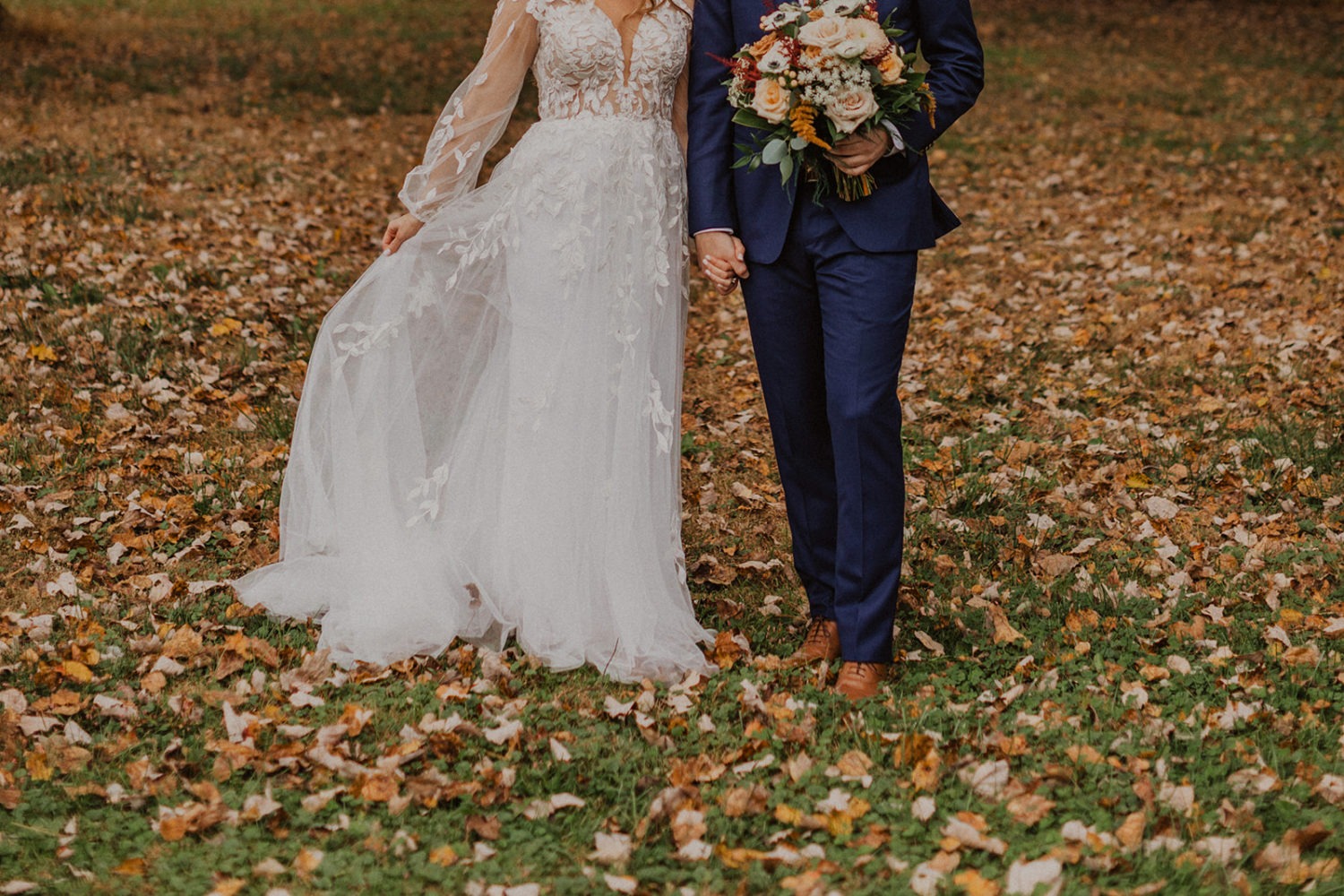 Bride holds dress and groom holds bouquet at fall leaves wedding