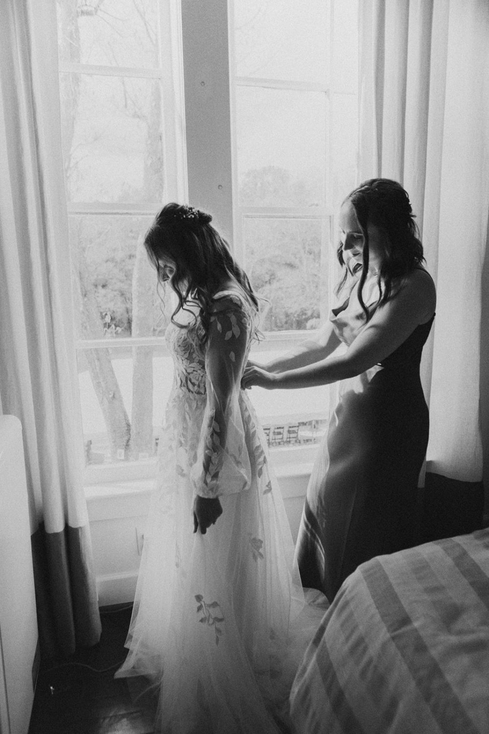 Bridesmaid zips up bride's wedding dress by window