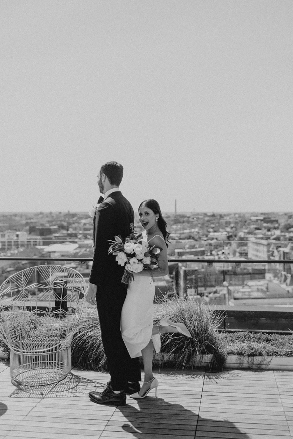Bride hugs groom from behind holding wedding bouquet on rooftop 