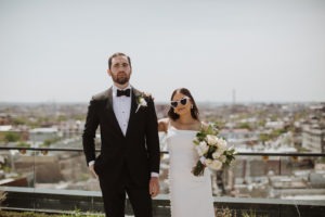 Couple poses with wedding bouquet and sunglasses at rooftop elopement