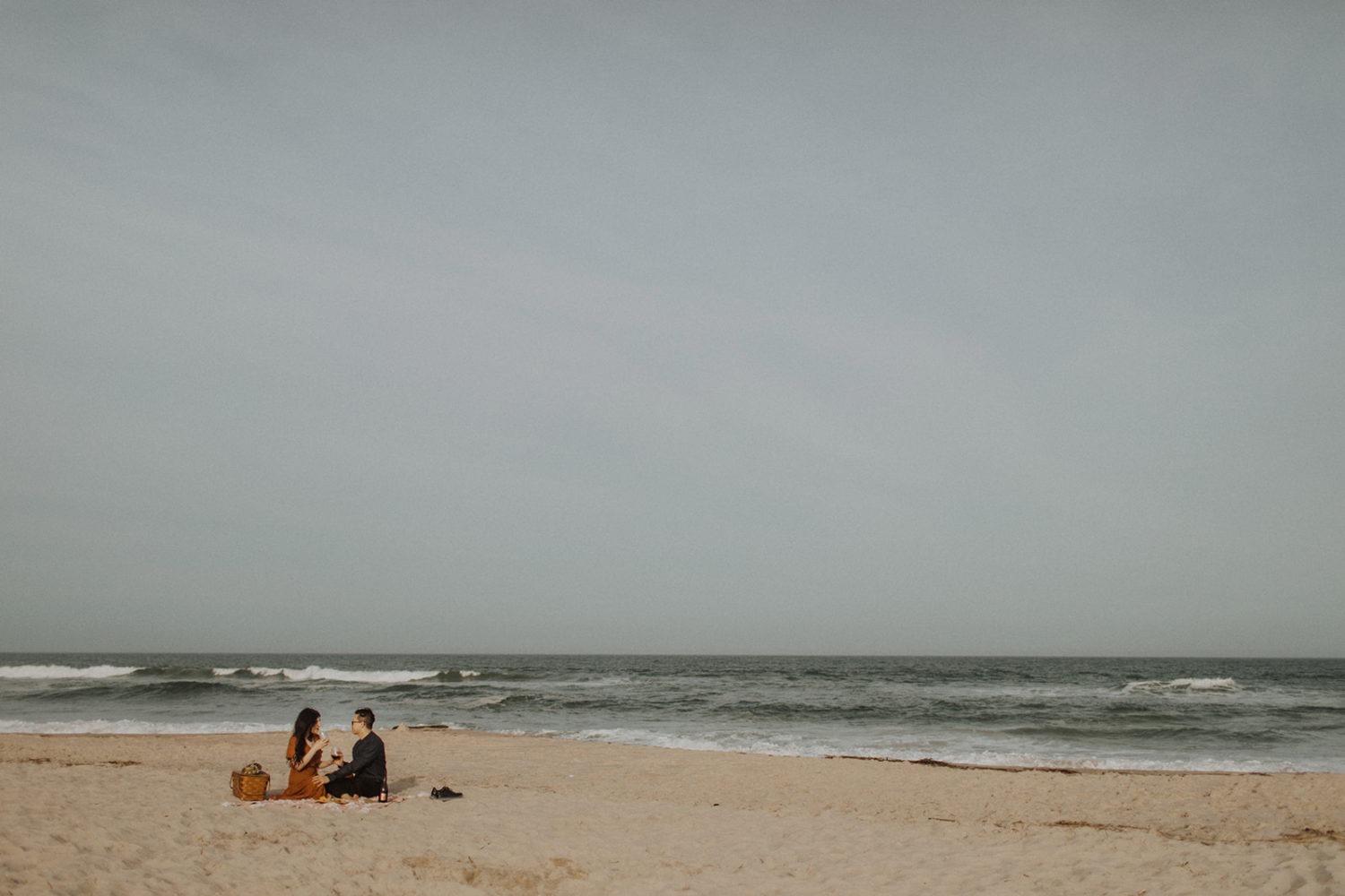 Couple sits on blanket with picnic basket for beach engagement photos