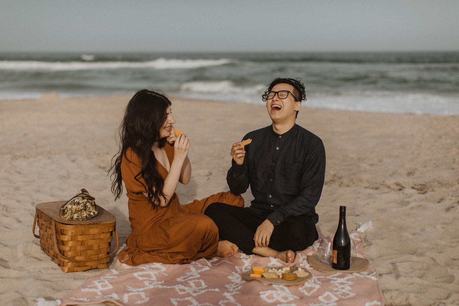 Couple eats on blanket with picnic basket for beach engagement photos
