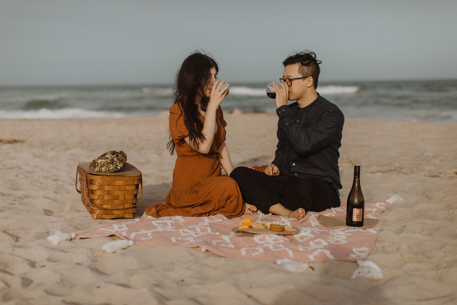 Couple drinks wine picnic blanket at beach engagement session