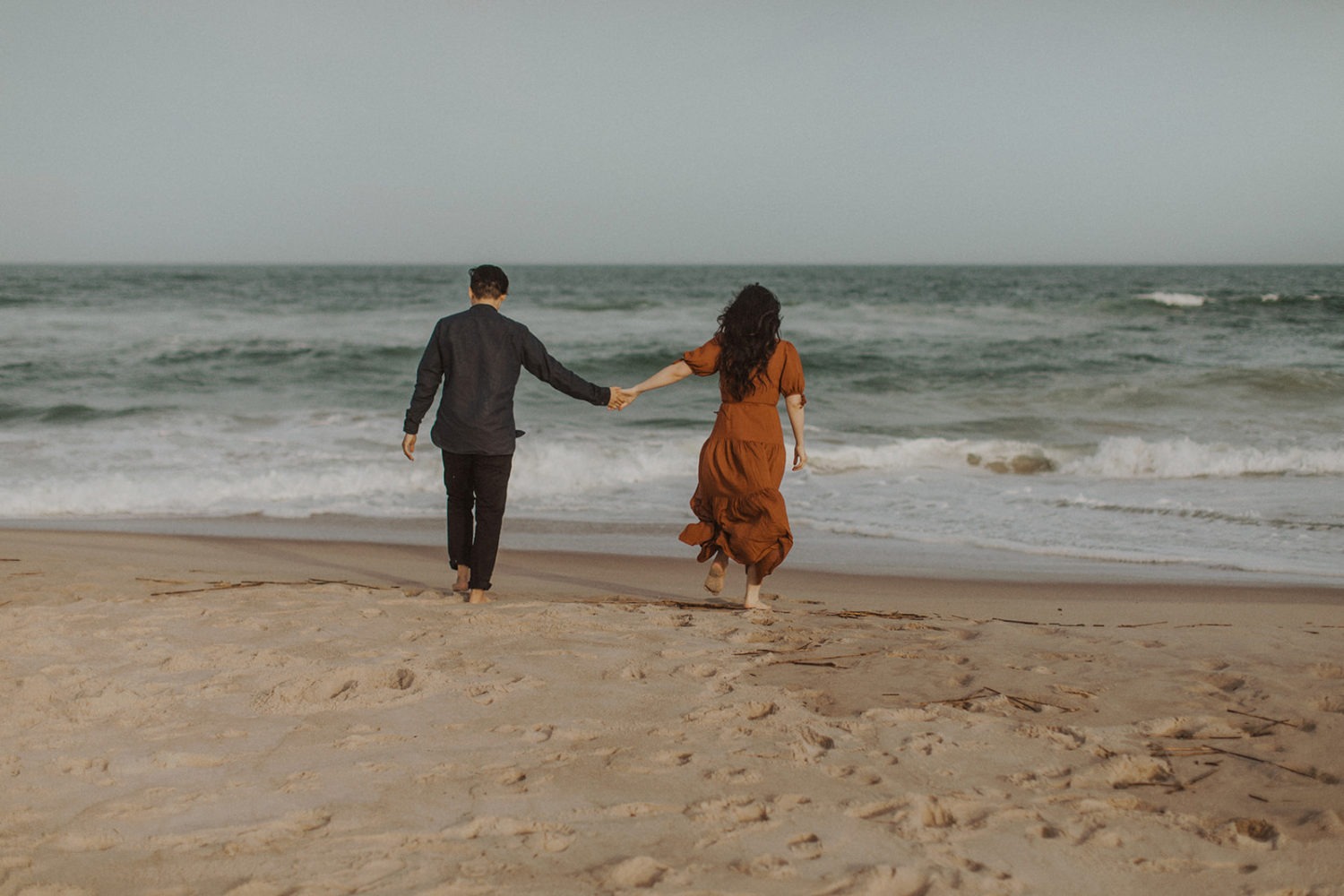 Couple walks towards ocean at beach engagement session