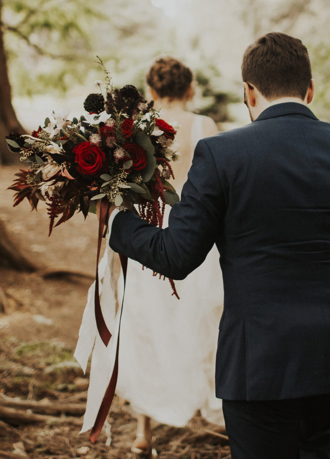 Groom holds bride's bouquet following her in woods first look