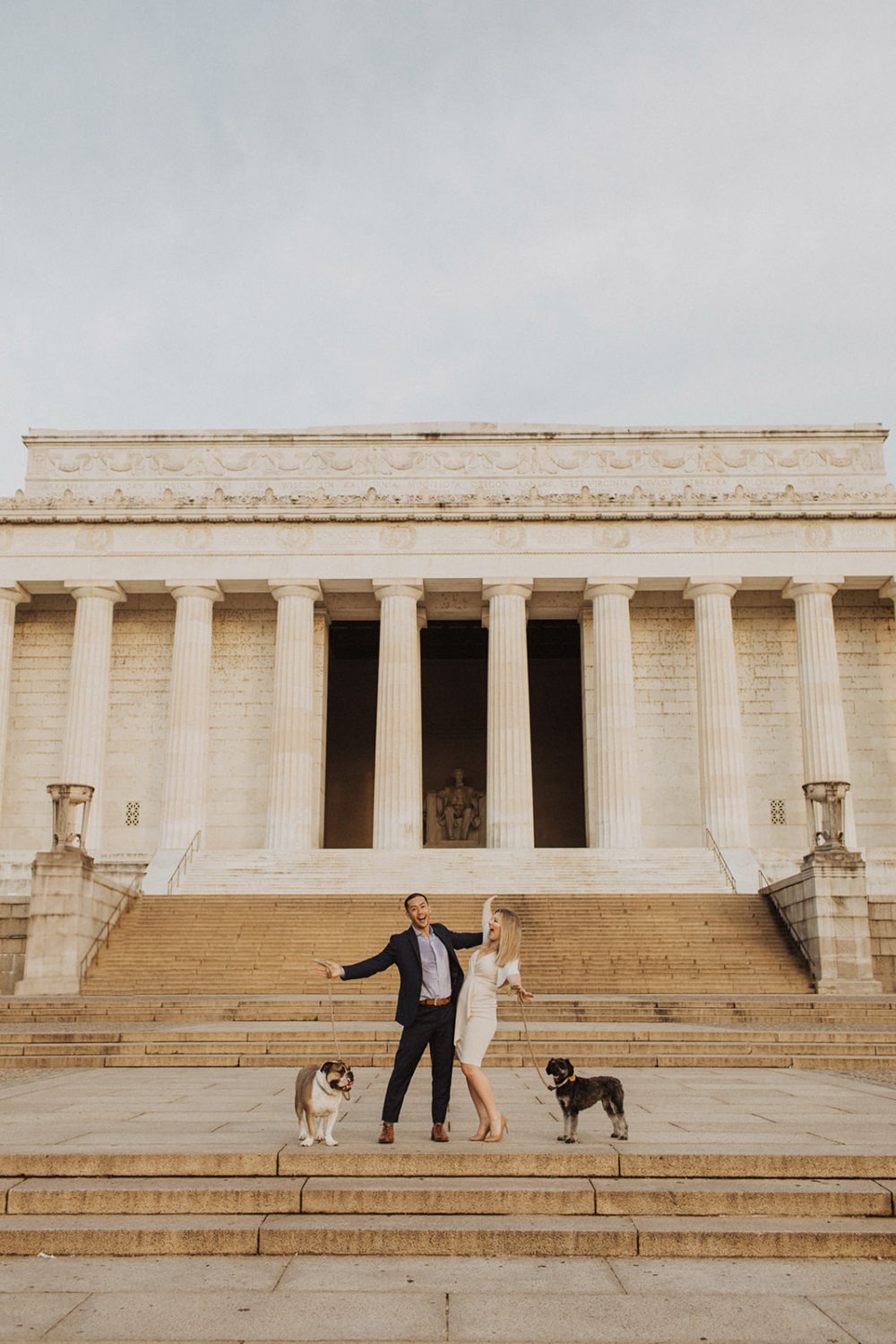 Couple stands with dogs in front of Lincoln Memorial at sunrise engagement session