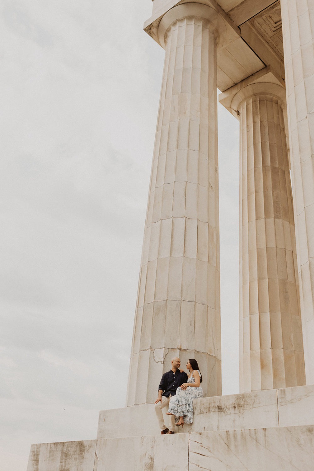 Couple sits on steps beside Lincoln Memorial columns at sunrise engagement session