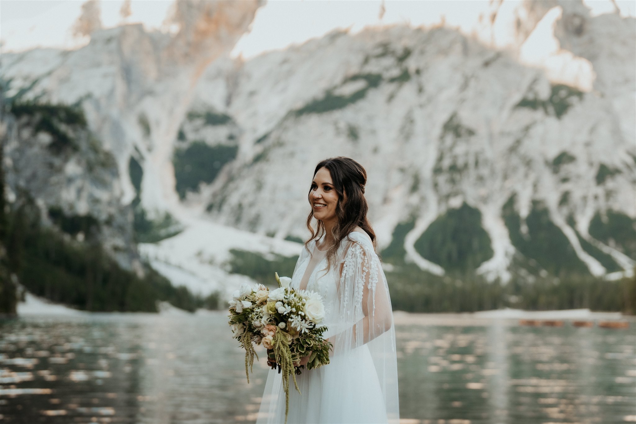 destination wedding photographer wears wedding dress and holds bouquet by lake