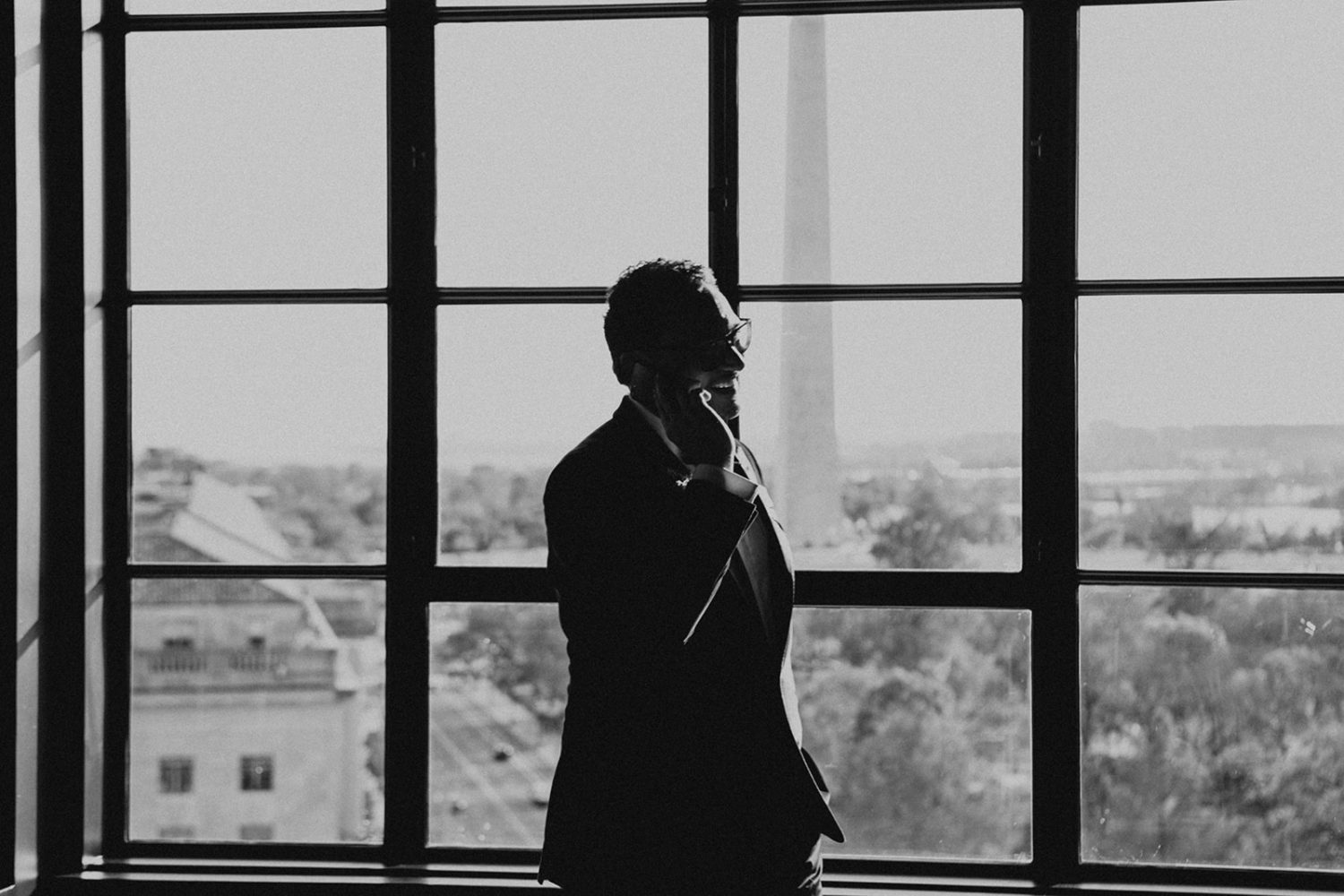 Groom chats on phone while in front of window onlooking the Washington Monument