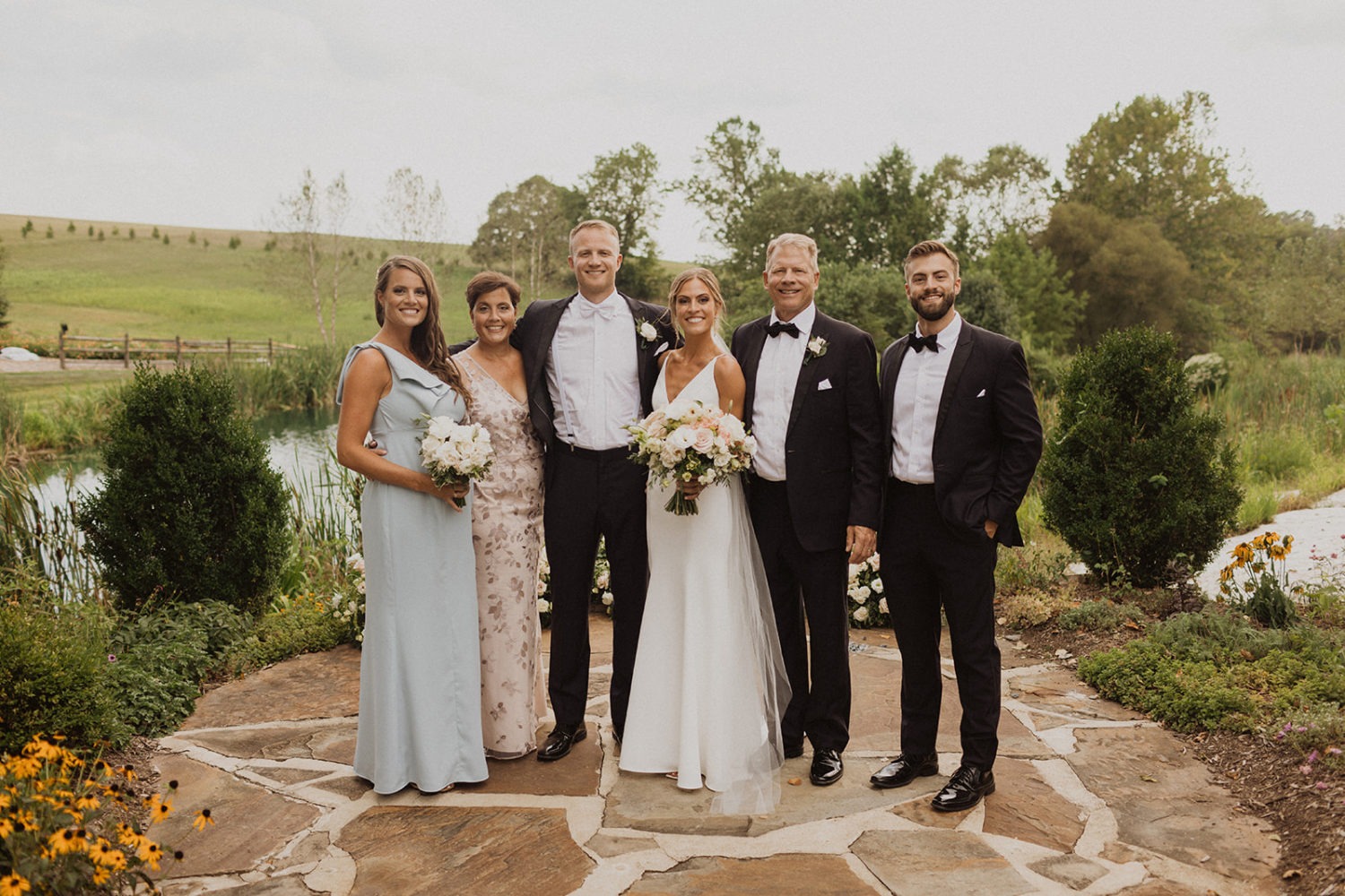 Couple stands with family by pond at farm wedding