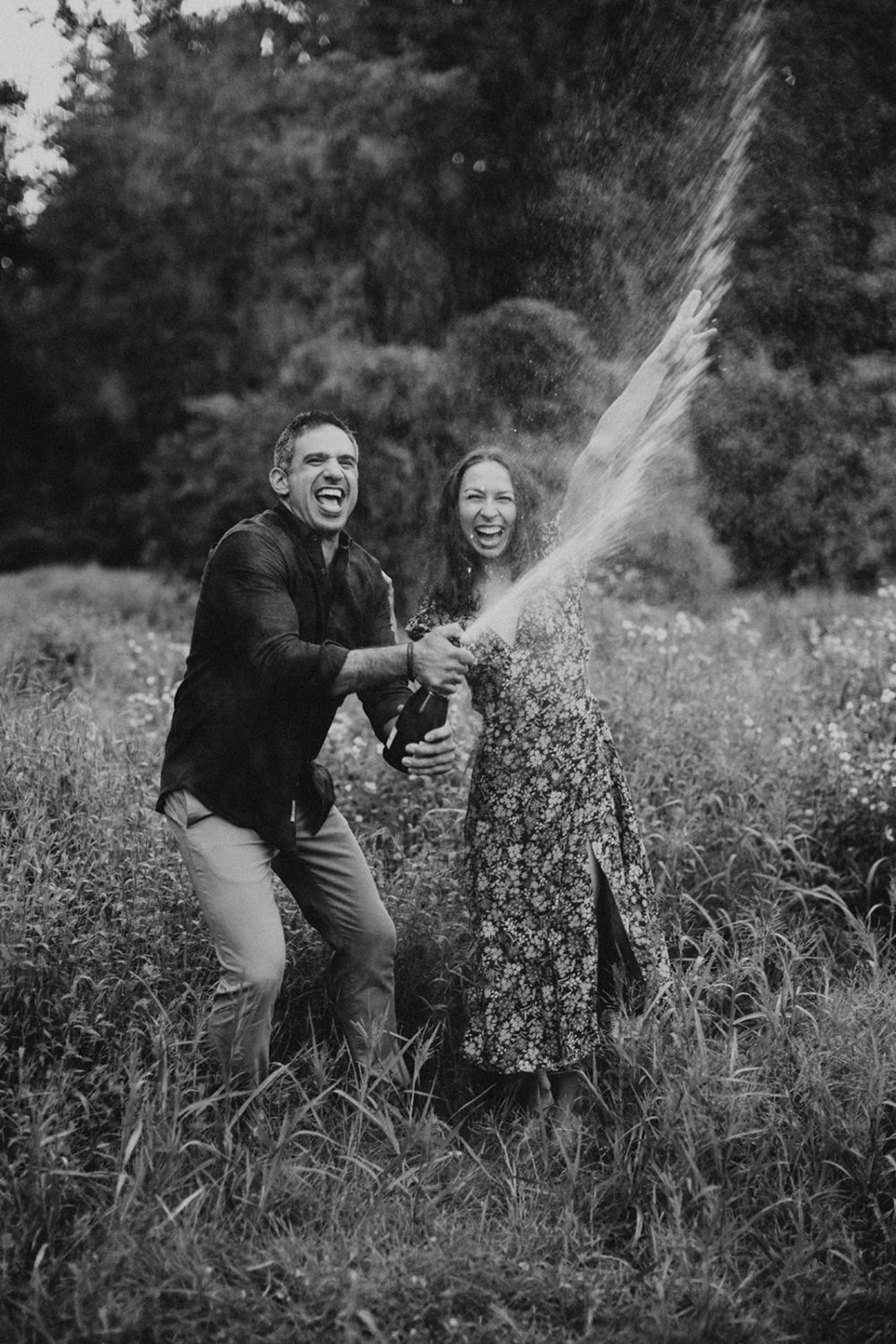 couple opens champagne bottle in field engagement session