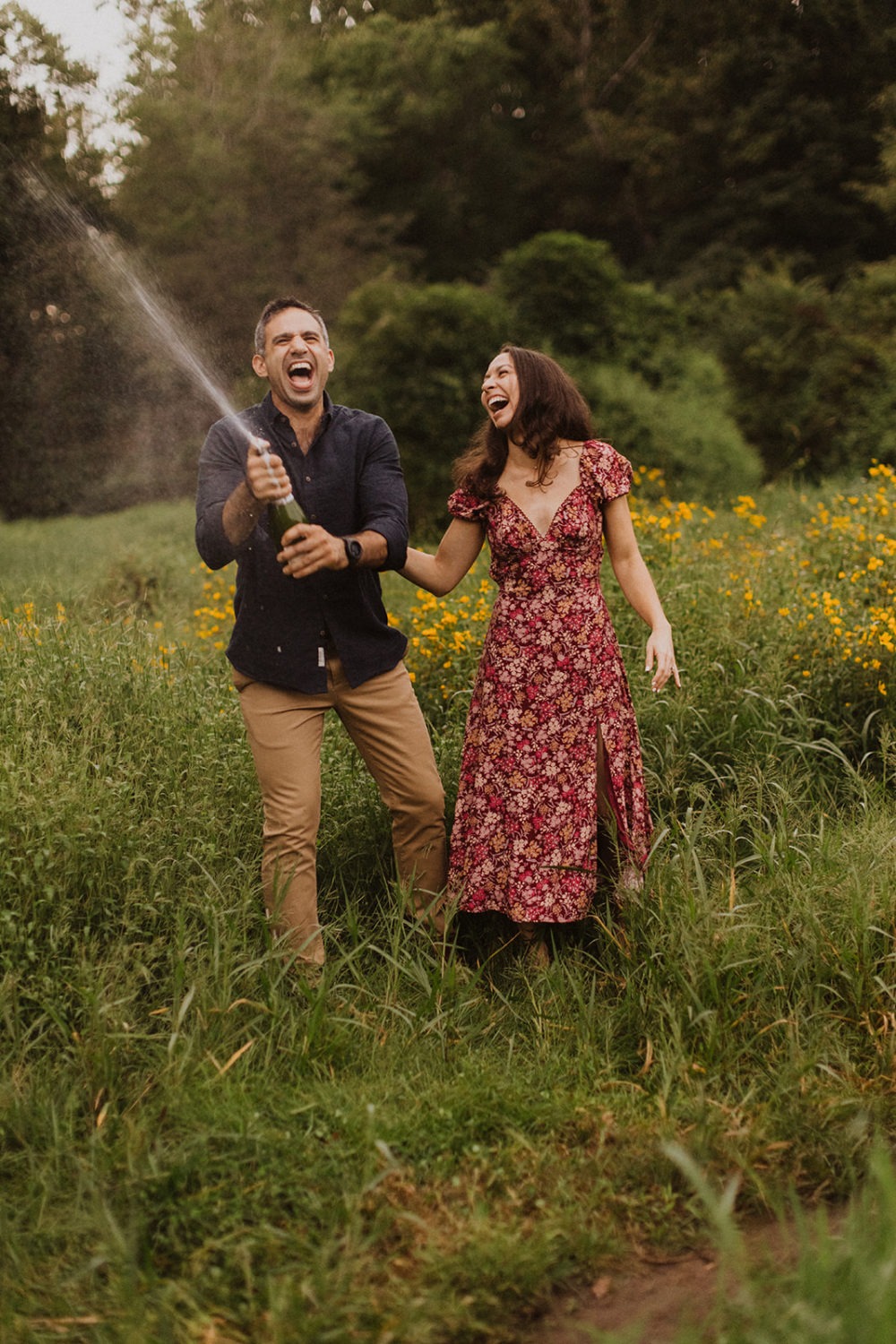 couple opens champagne bottle in field engagement session