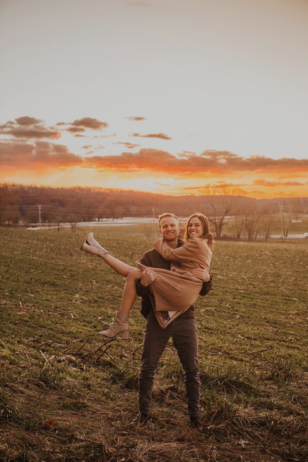 man lifts woman in field at sunset
