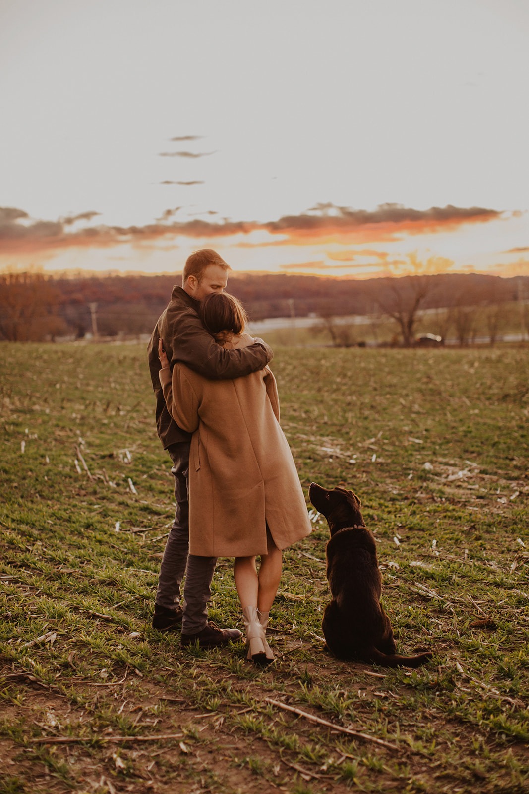 Couple stands in field at sunset fall engagement session with dog