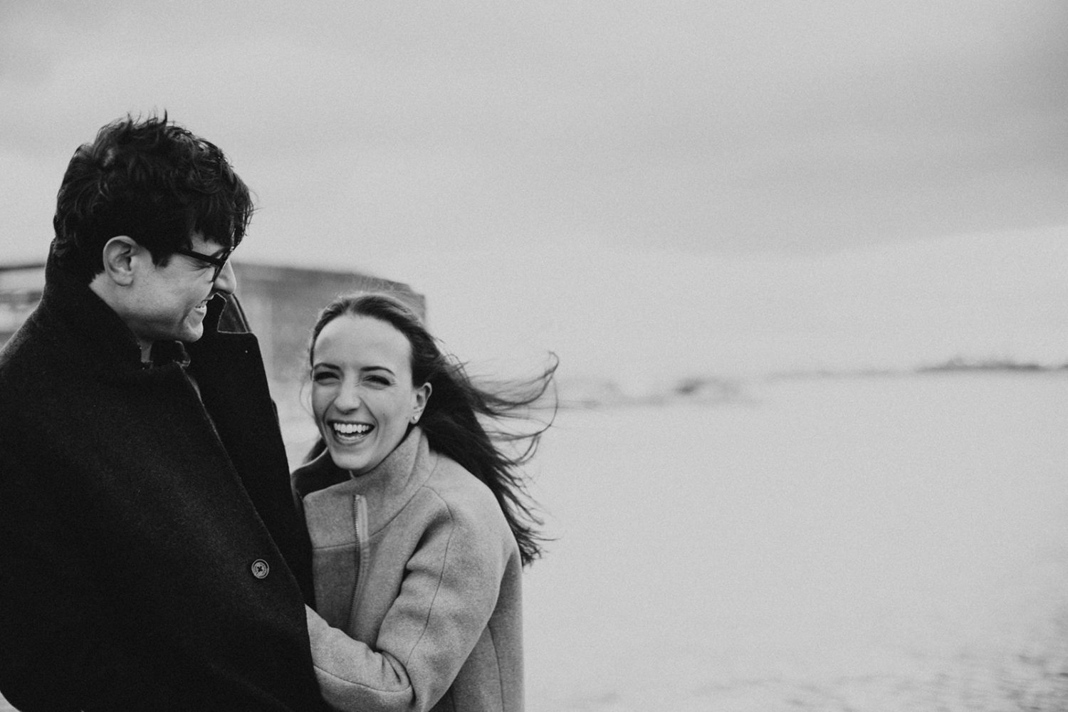 couple laughs together at windy winter engagement session
