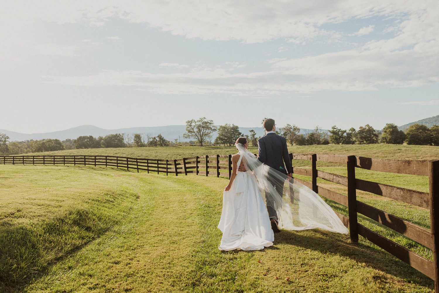 couple walks holding hands along wooden fence in virginia field