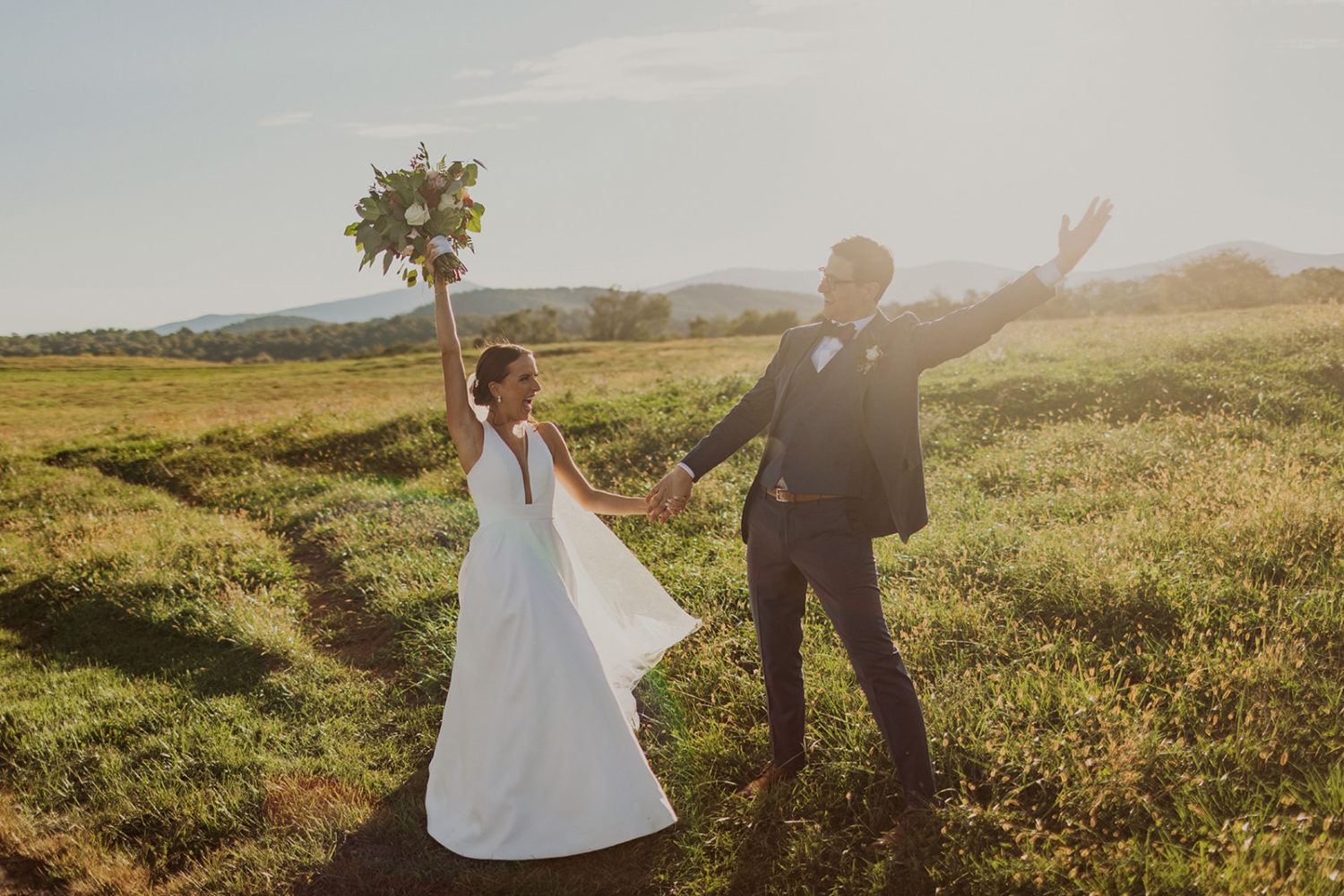 couple raises arms cheering in field at sunset wedding