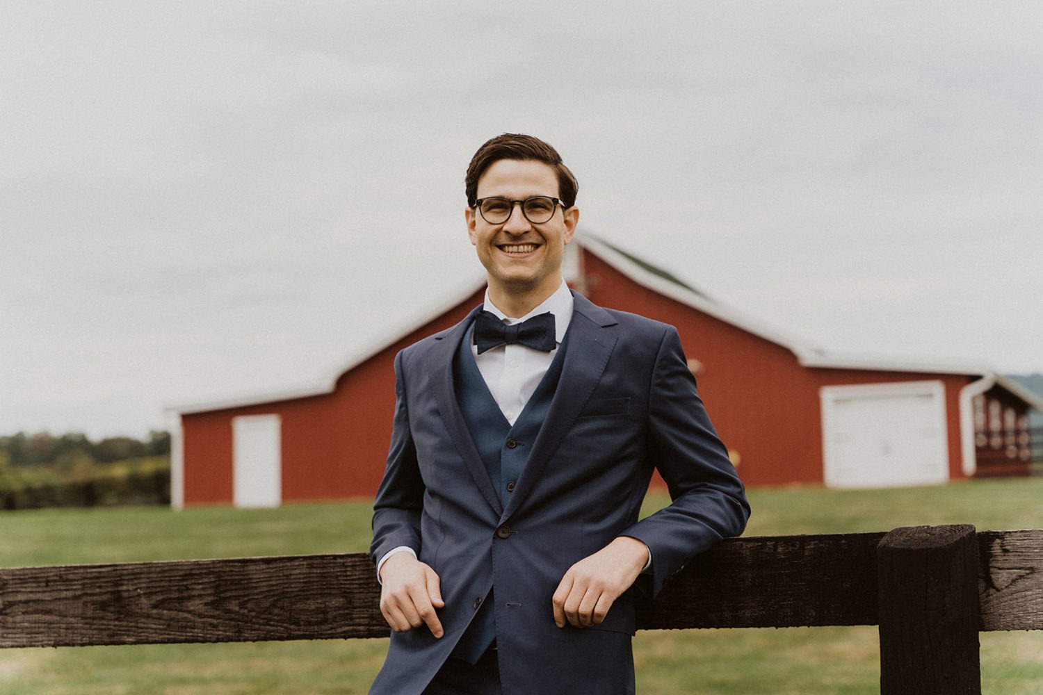 groom leans on wooden fence in front of stables at wedding ranch venue