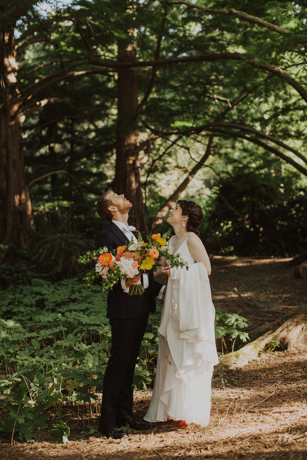 wedding couple looks up at trees holding bouquet at garden wedding