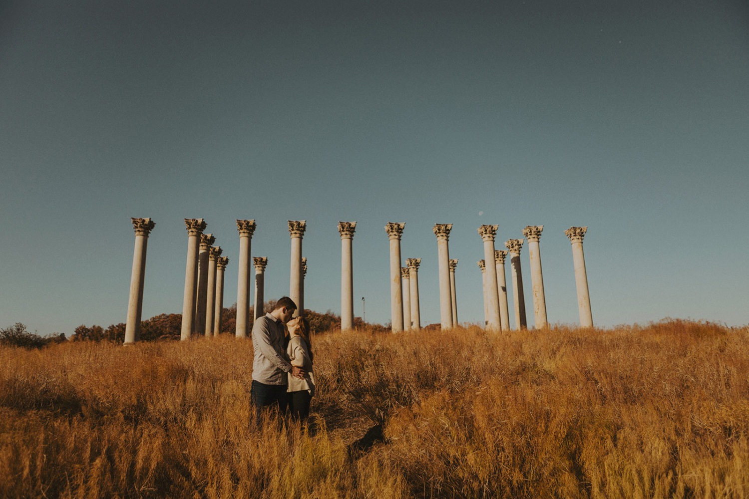 couple stands in field in front of National Columns at engagement session