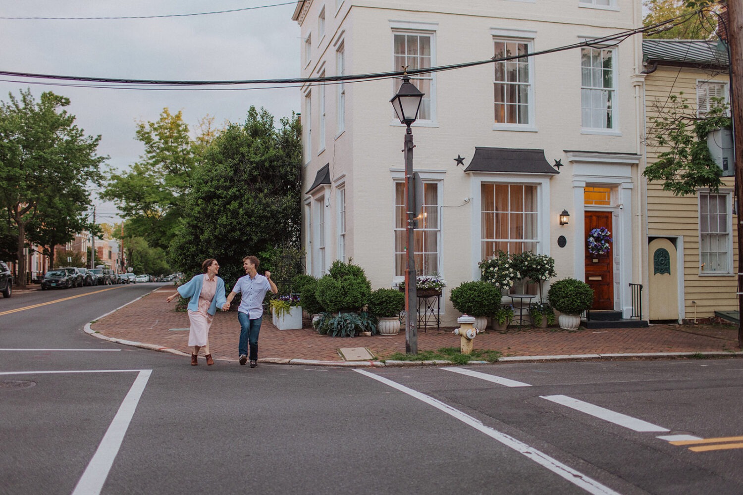 couple crosses street holding hands at Old Town Alexandria waterfront engagement session