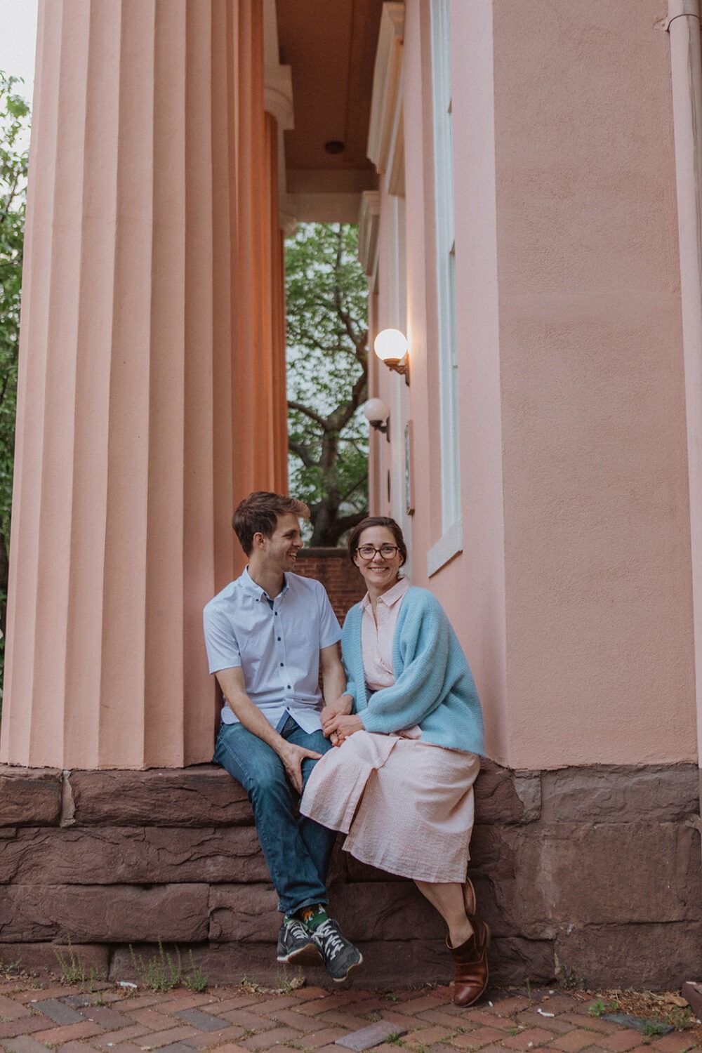 couple sits in front of pink column building at engagement session