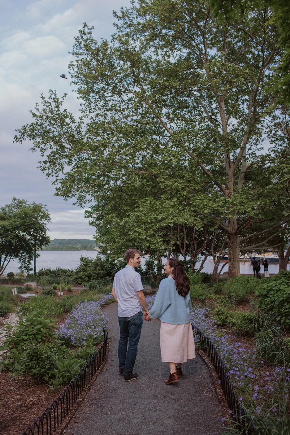couple walks holding hands through park at Old Town Alexandria waterfront engagement session