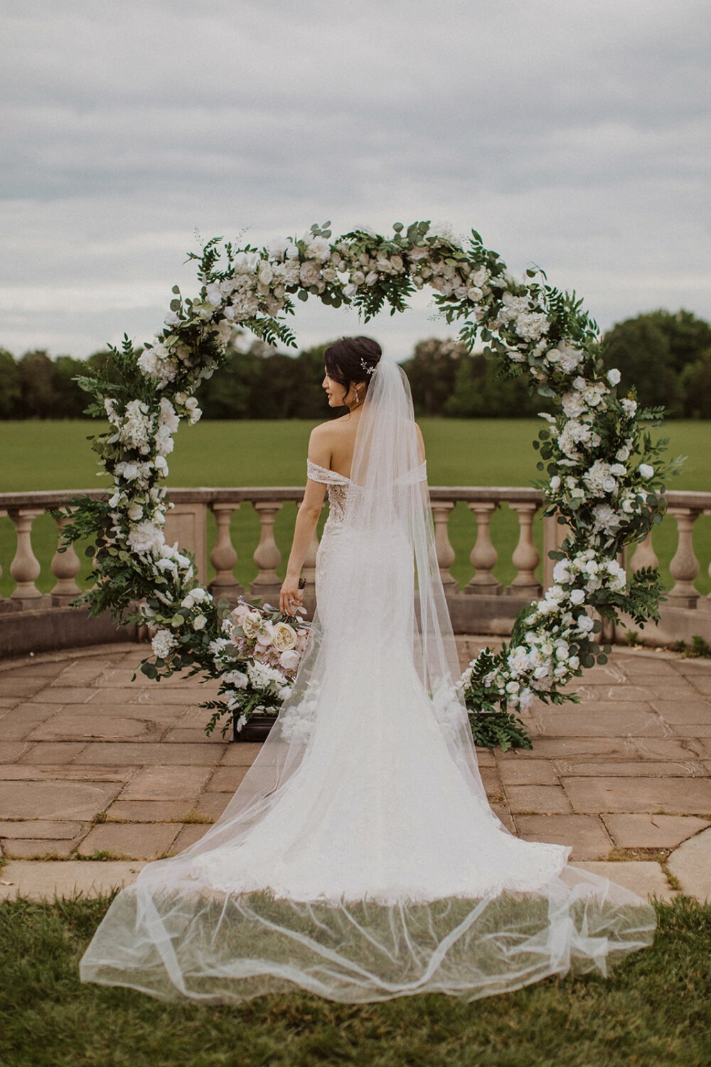 bride stands holding bouquet under white floral wedding arch