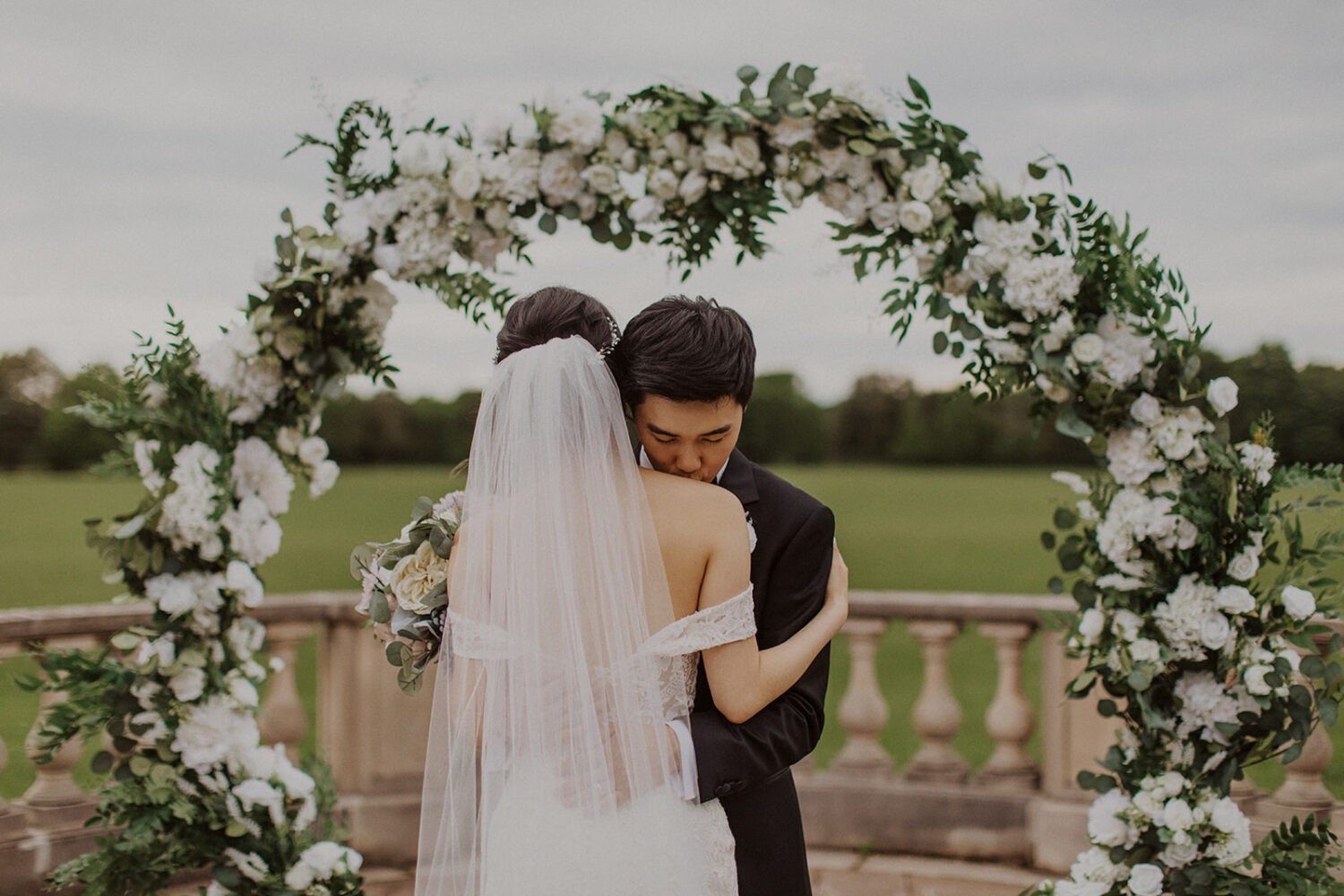 groom embraces bride while standing under white floral arch