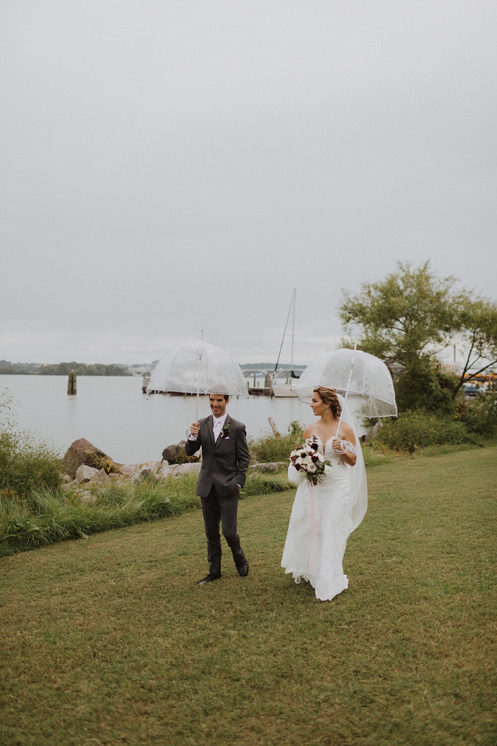 couple walk along waterside holding clear umbrellas