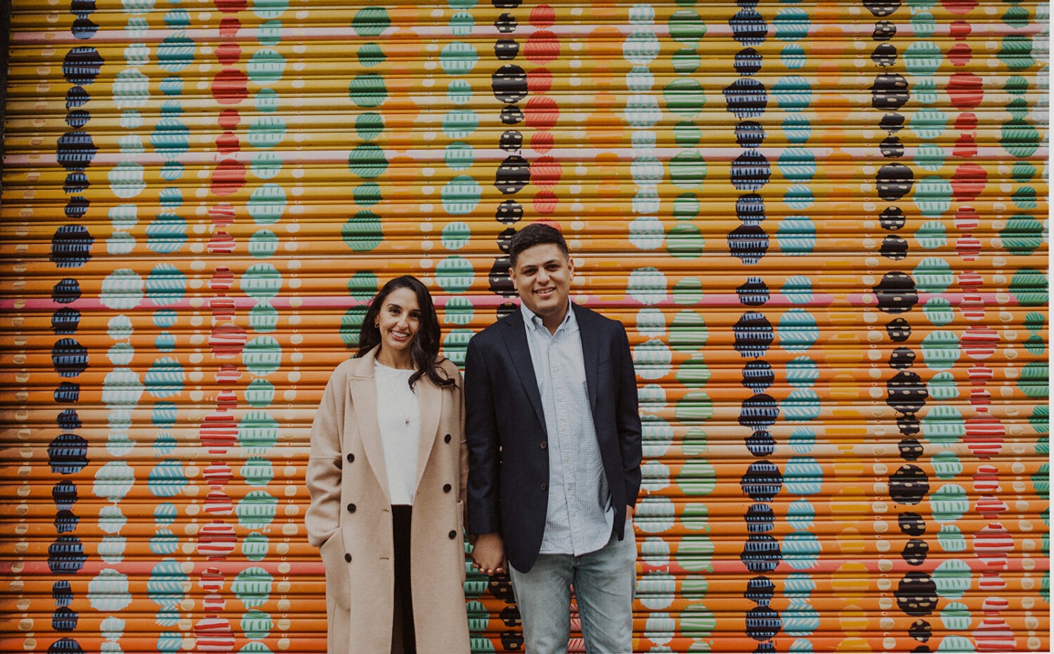 couple stands holding hands in front of rainbow-colored wall