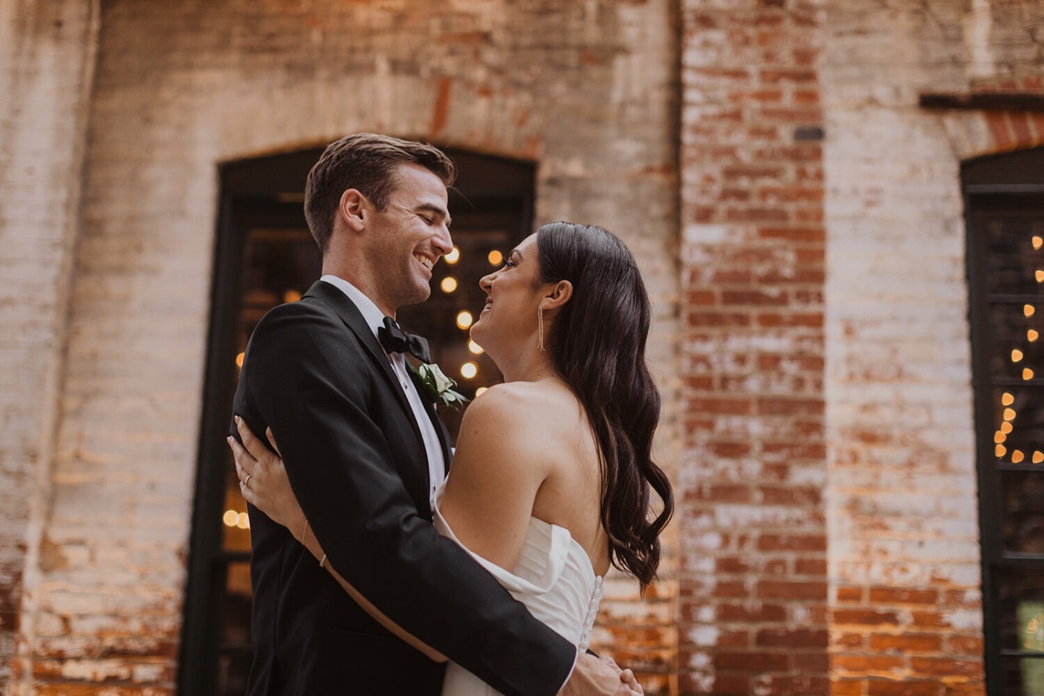 couple embraces in front of white brick wall at Mt. Washington Mill Dye House wedding venue