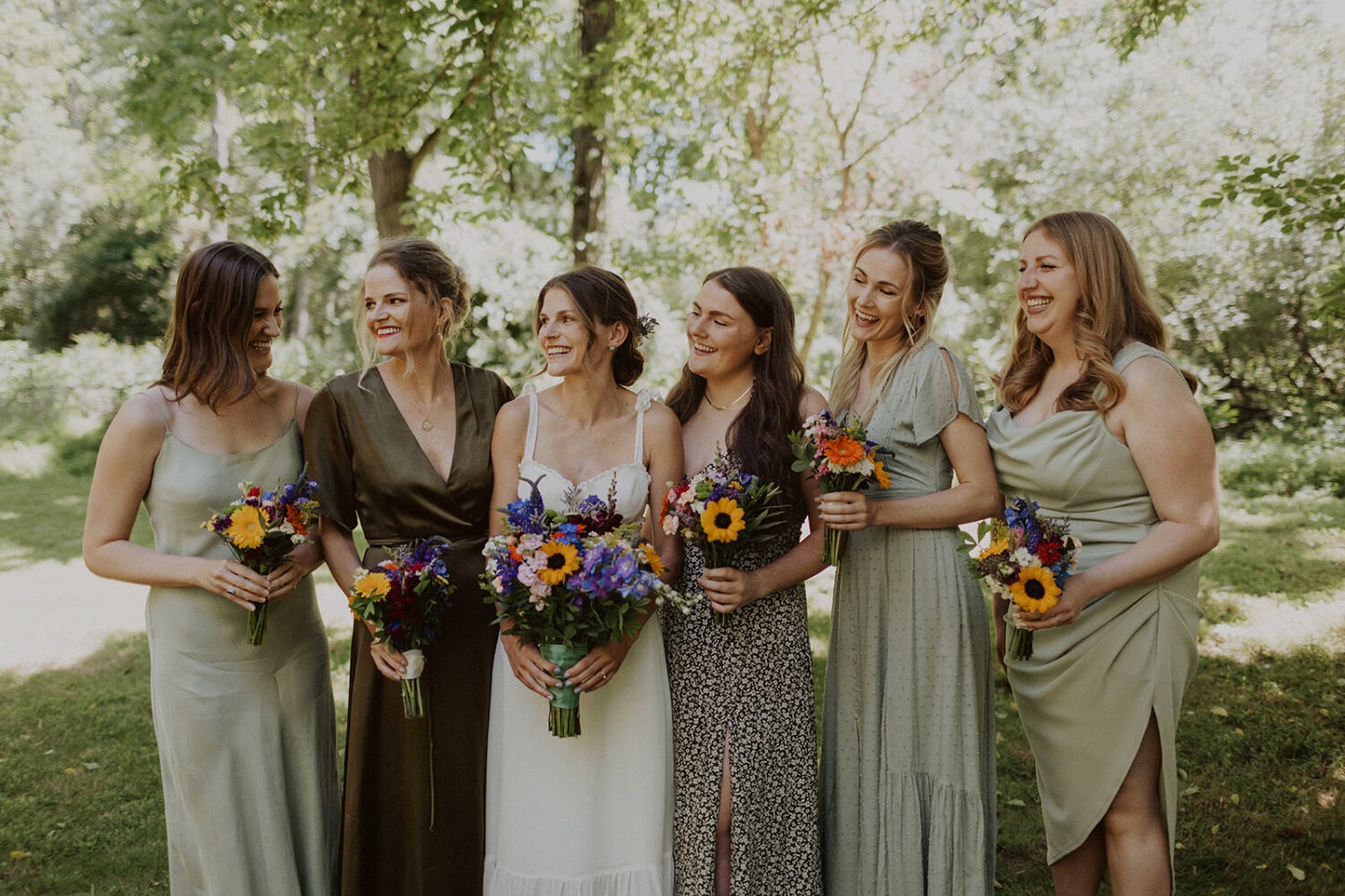 bride laughs with bridesmaids holding wildflower bouquets at rustic wedding
