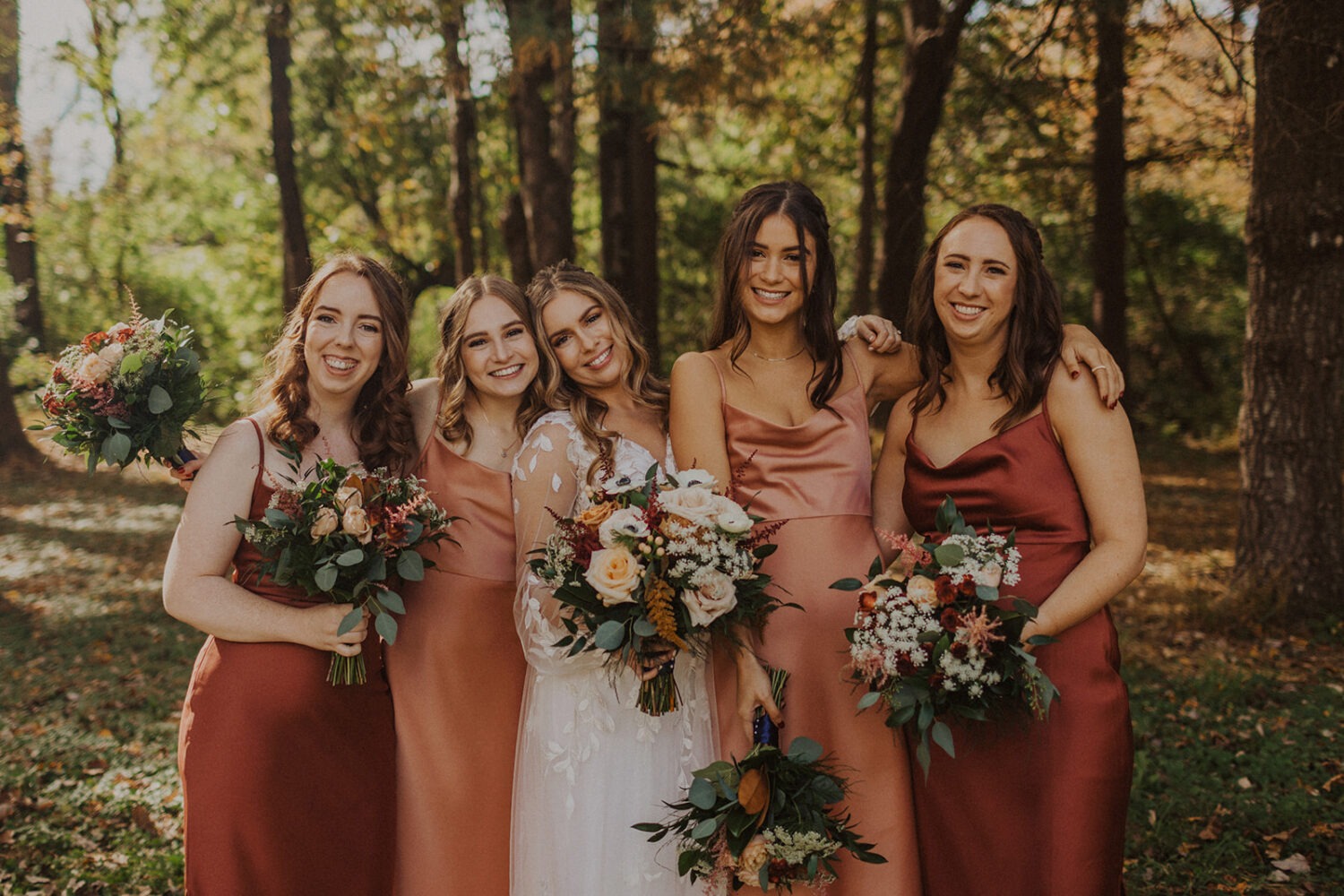 bridesmaids embrace with bride holding bouquets at outdoor wedding 