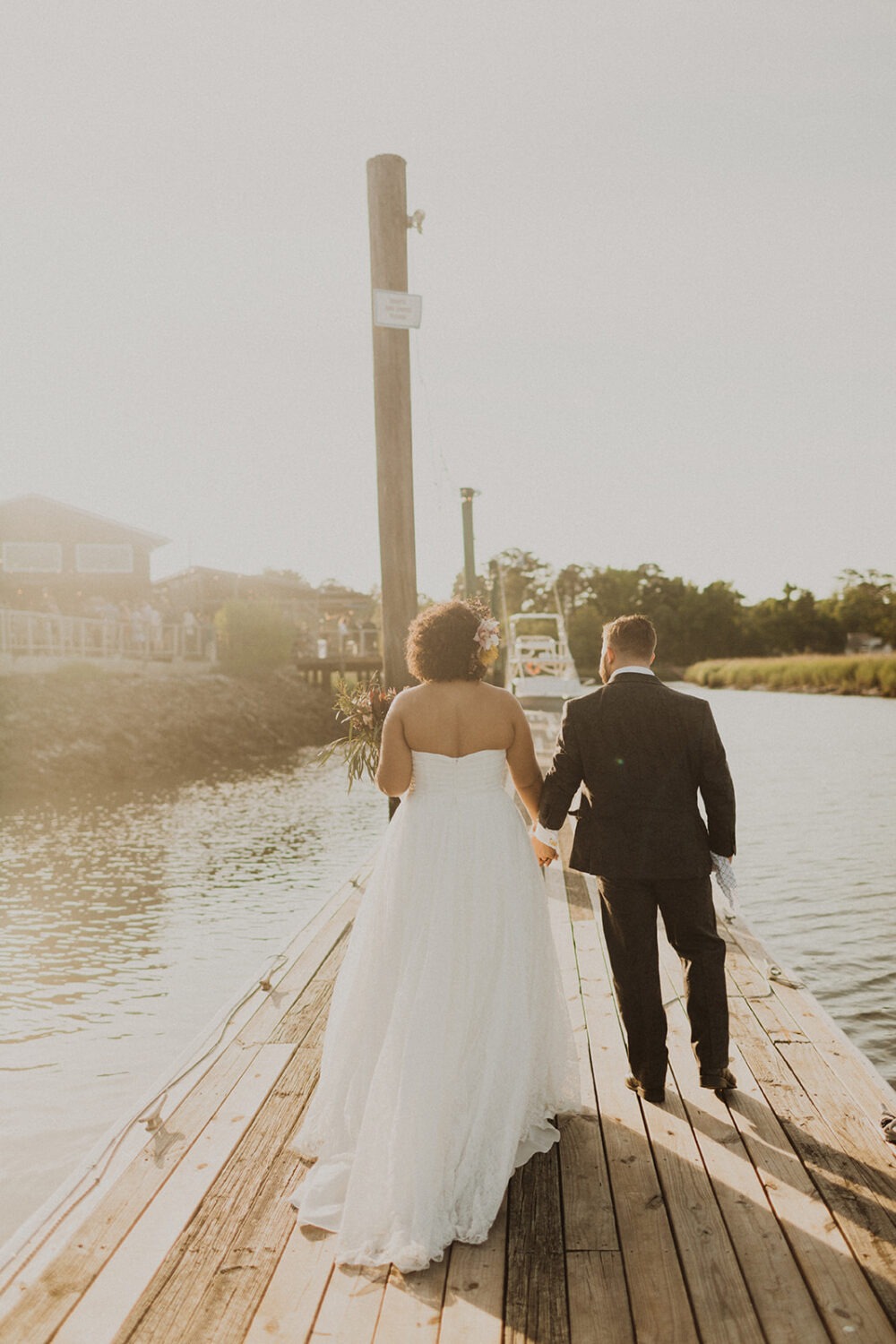 couple holds hands walking along dock at summer wedding waterfront