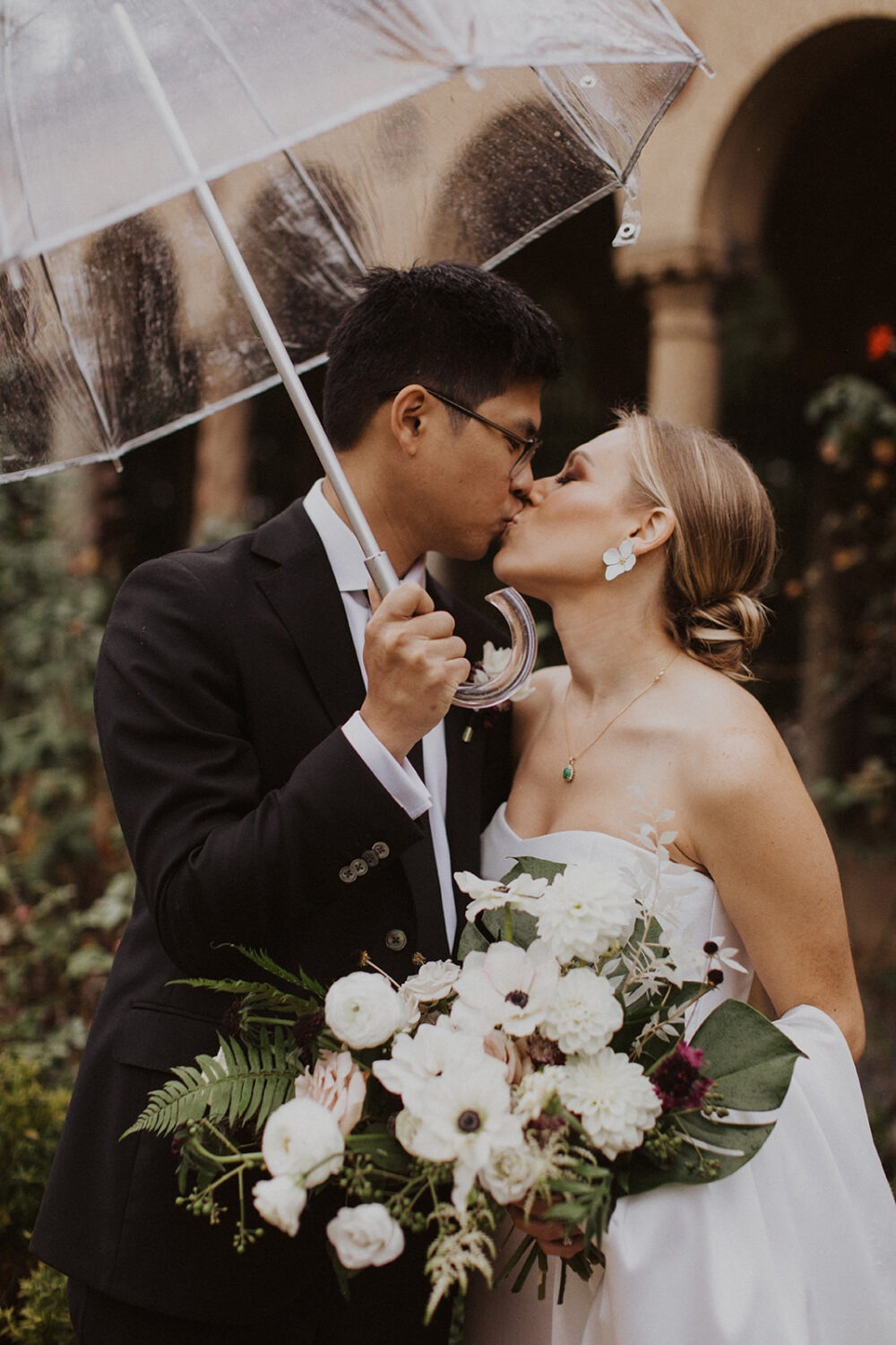 couple kisses under clear umbrella at rainy wedding