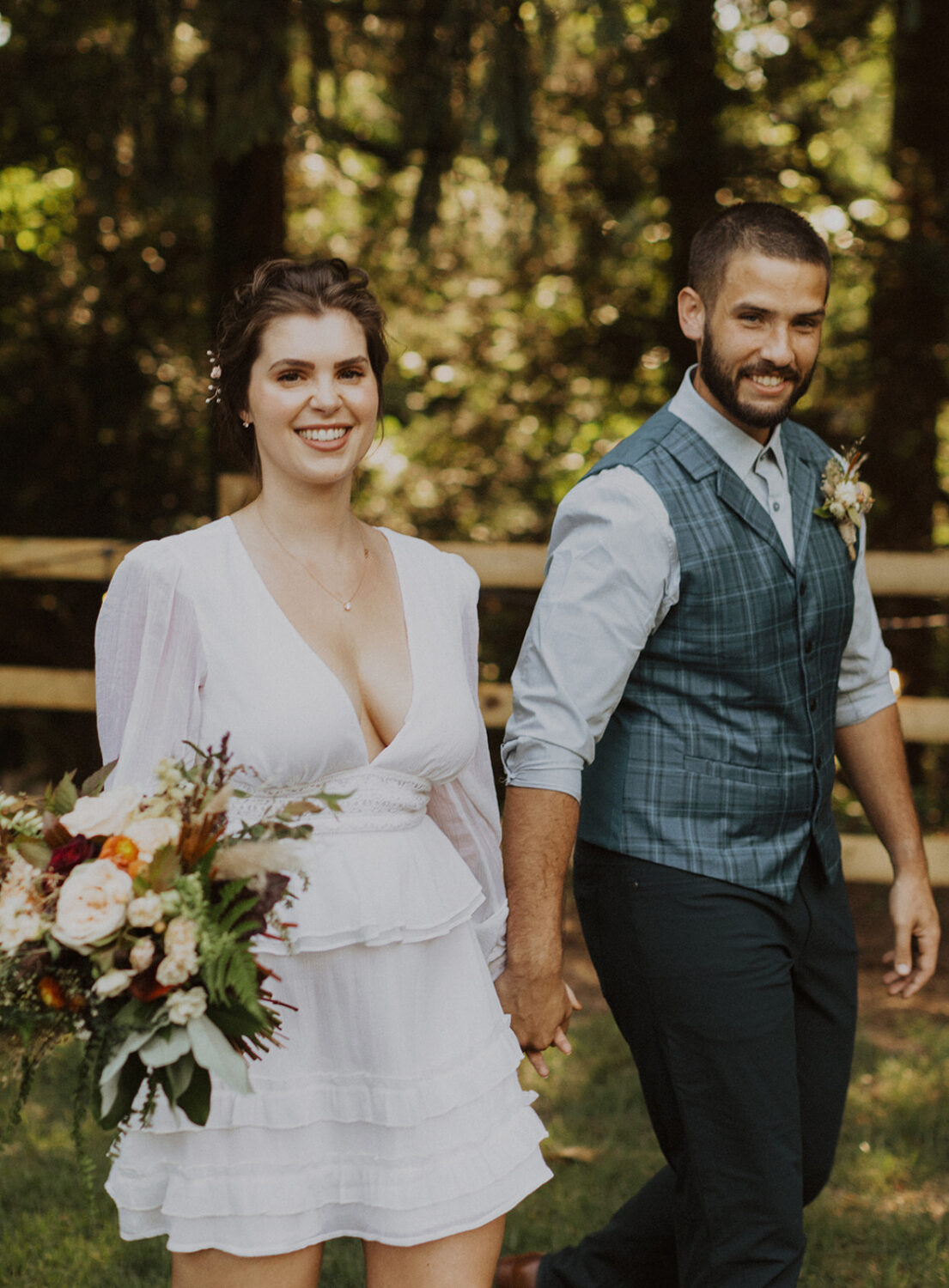 couple walks holding hands and bouquet in backyard 
