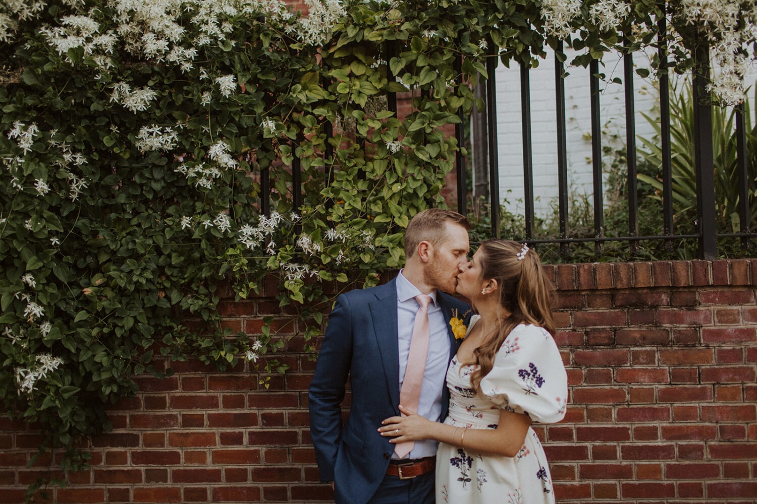 couple kisses in front of brick and flowers at Dumbarton House garden wedding