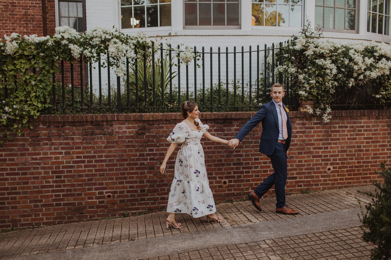 couple walks holding hands in front of brick and flowers at Dumbarton House garden wedding