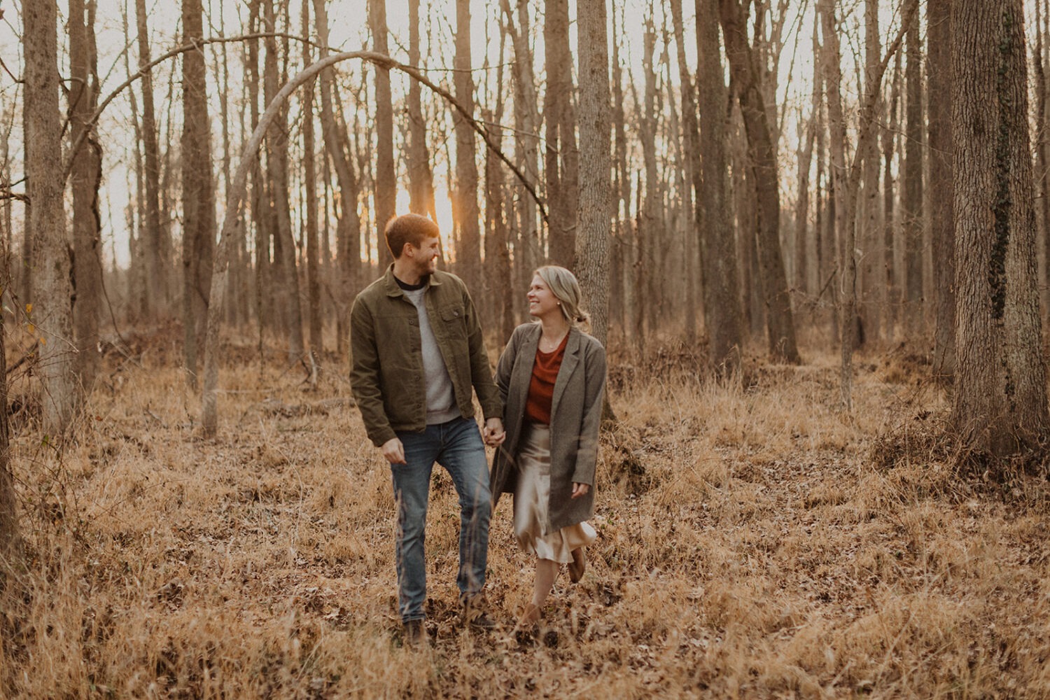 couple holds hands walking in forest at sunset engagement session