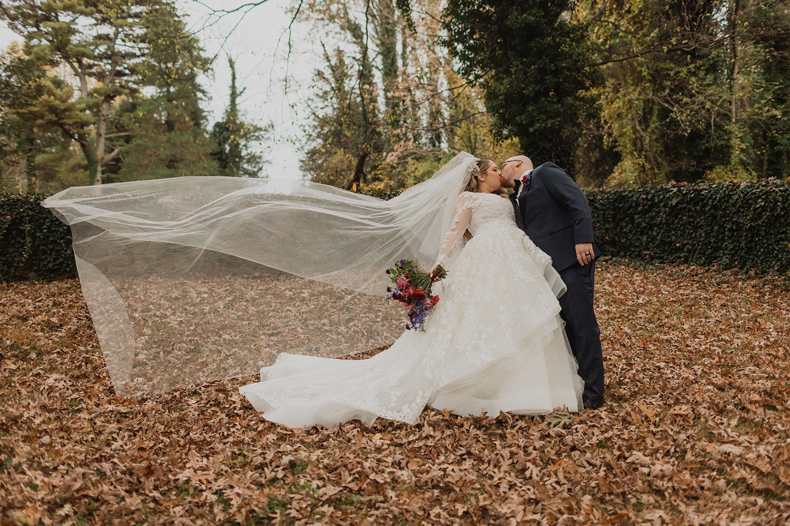 couple kisses with veil in wind at fall outdoor wedding