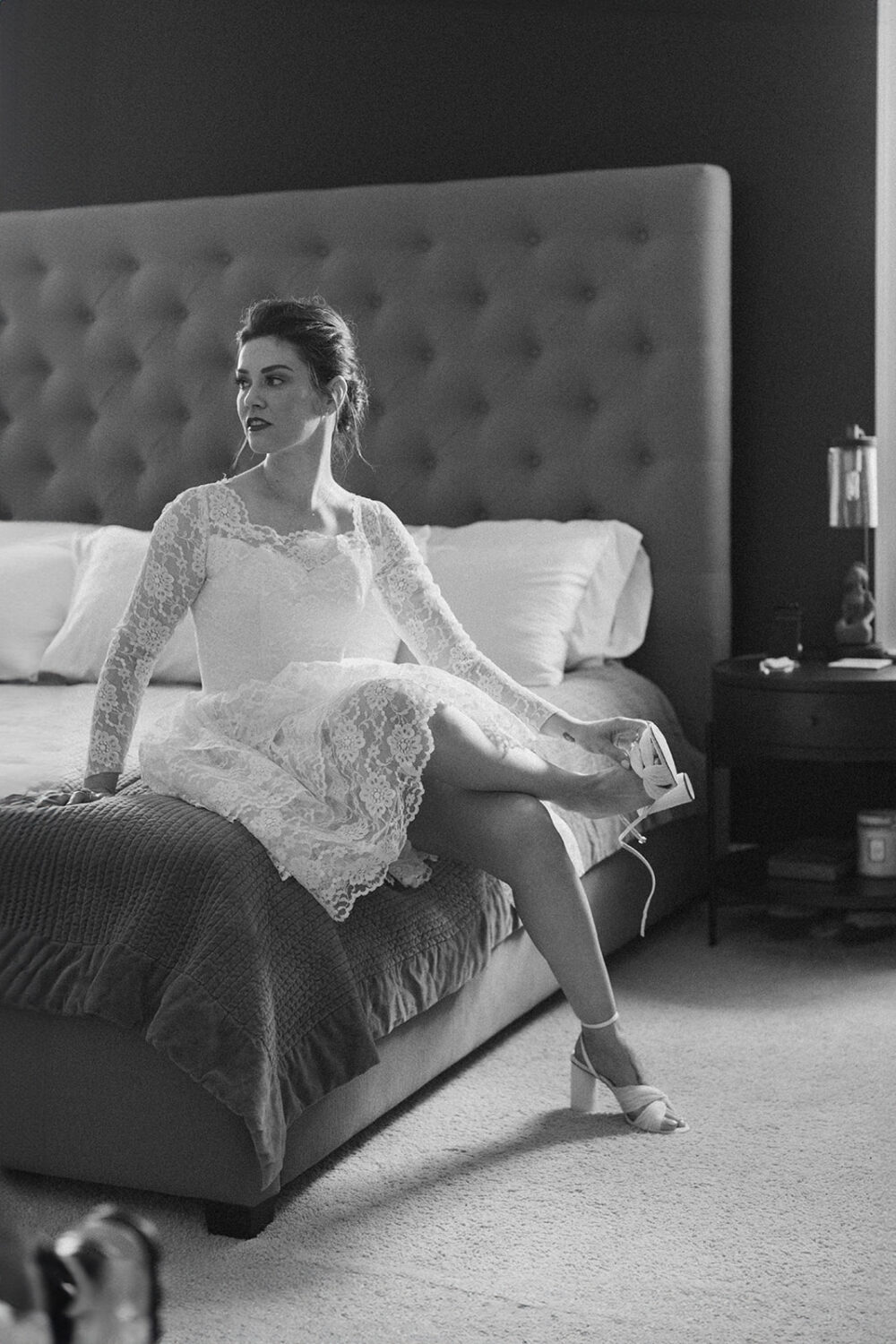 bride sits on bed putting on wedding shoes