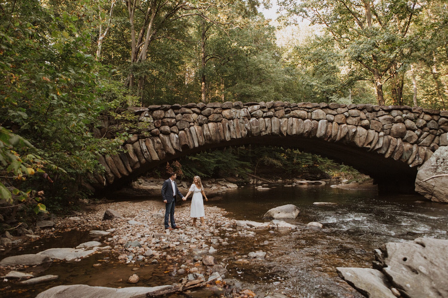 couple walks along creek under bridge for Rock Creek Park photos