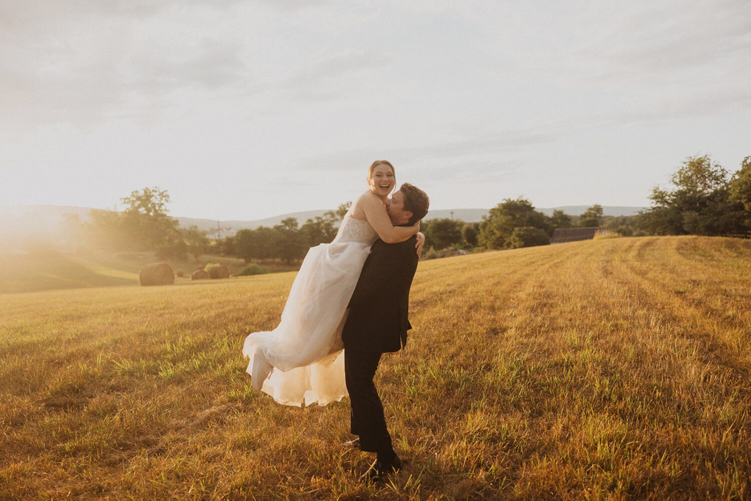 groom carries bride at sunset outdoor field wedding venue