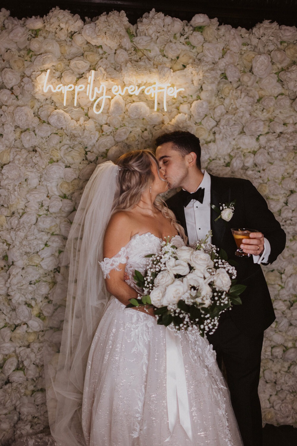 couple kisses under neon sign and wall of white roses