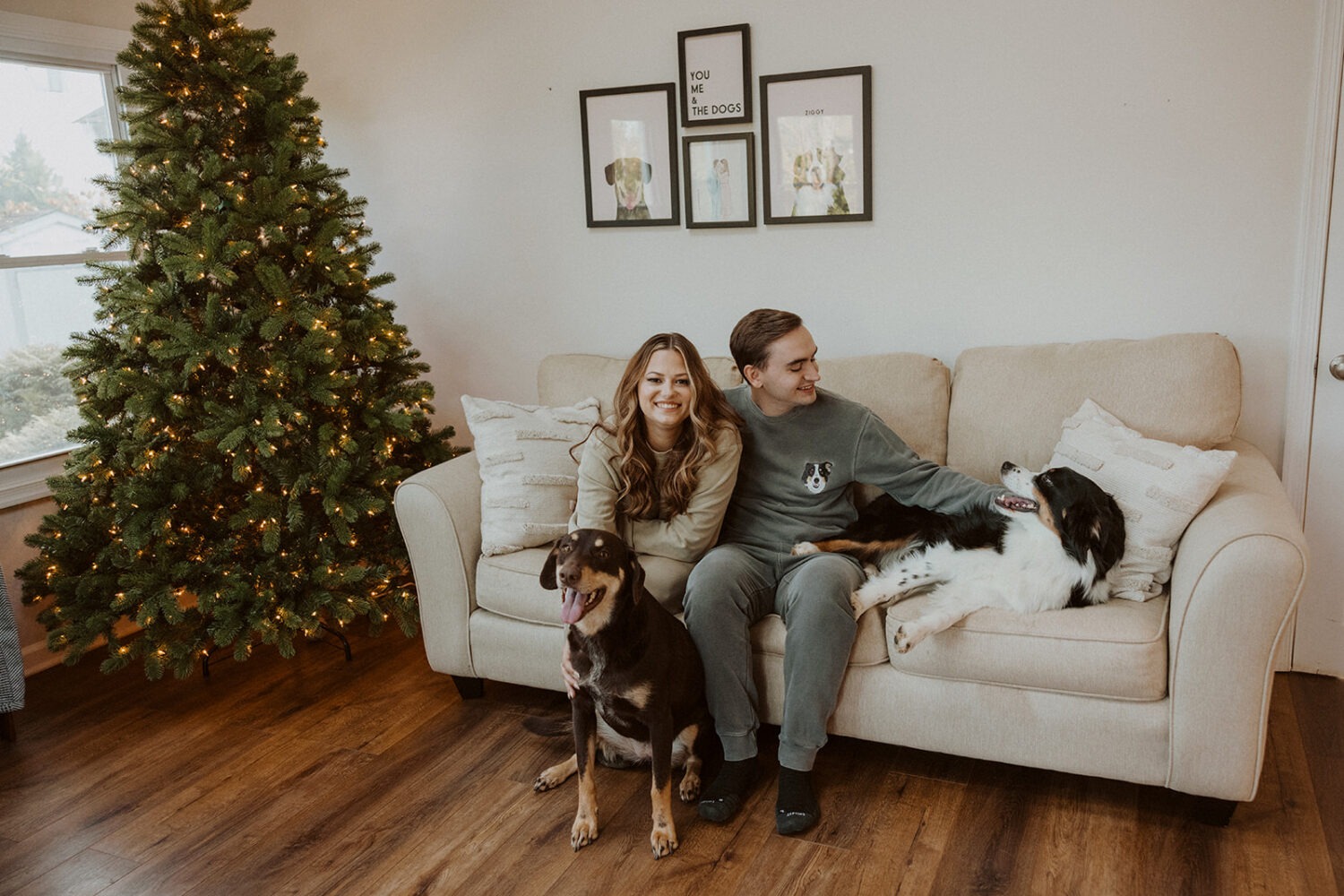 couple sits with dogs in front of Christmas tree at winter engagement photoshoot